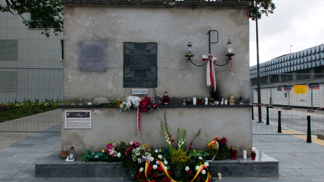 Fragment przedwojennego muru Zieleniaka. Na nim tablice upamiętniające cywilne ofiary Powstania. Obok kotwica Polski walczącej. U podstawy ściany kwiaty i wieńce.