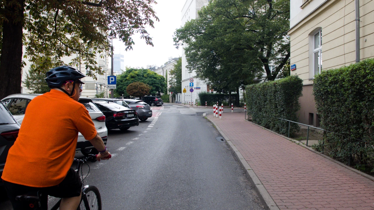 Na wąskiej ulicy rowerzysta. Po prawej chodnik i kamienice. Po lewej zaparkowane samochody.