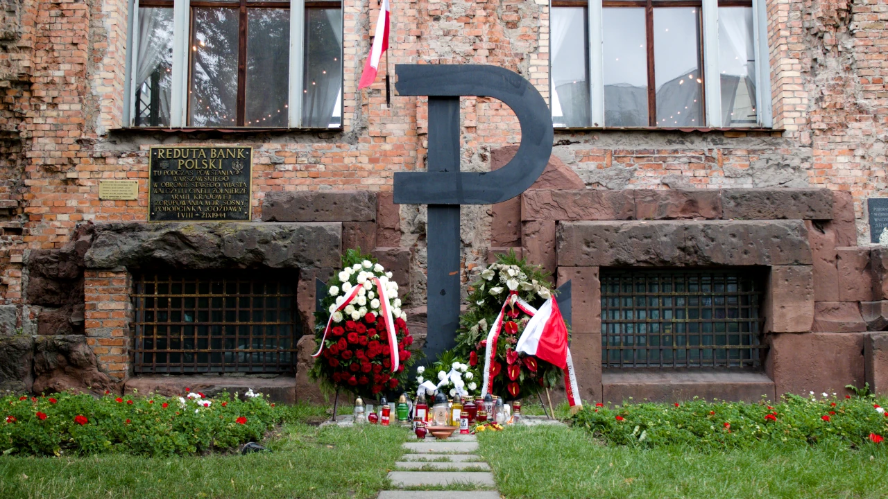 Ściana budynku Reduty Banku Polskiego. Na trawniku przed budynkiem stoi symbol Polski walczącej, sięgający do okien wysokiego parteru. Pod kotwicą widać wieńce i kwiaty.