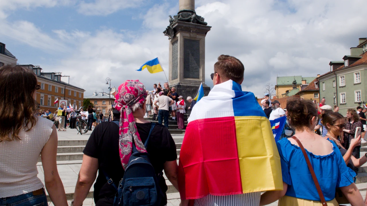 Widoczni od tyłu ludzie na manfestacji stojący w kręgu i trzymający się za ręce. Na pierwszym planie mężczyzna owinięty we flagę z łączącą barwy narodowe Polski i Ukrainy. W tle łopoce Ukraińska flaga.