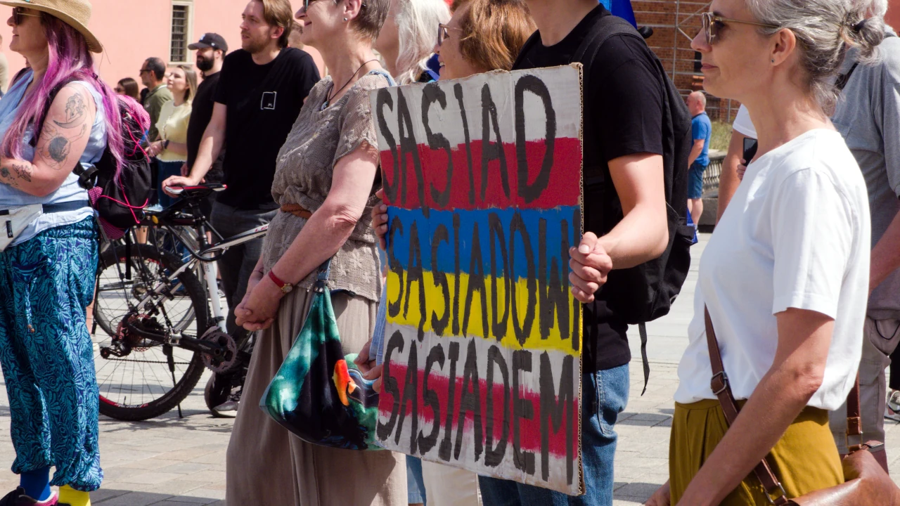 Kilka osób stojących na placu. Jedna osoba trzyma plakat z w barwach narodowych Polski, Ukrainy i Białorusi z napisem: sąsiad sąsiadowi sąsiadem.