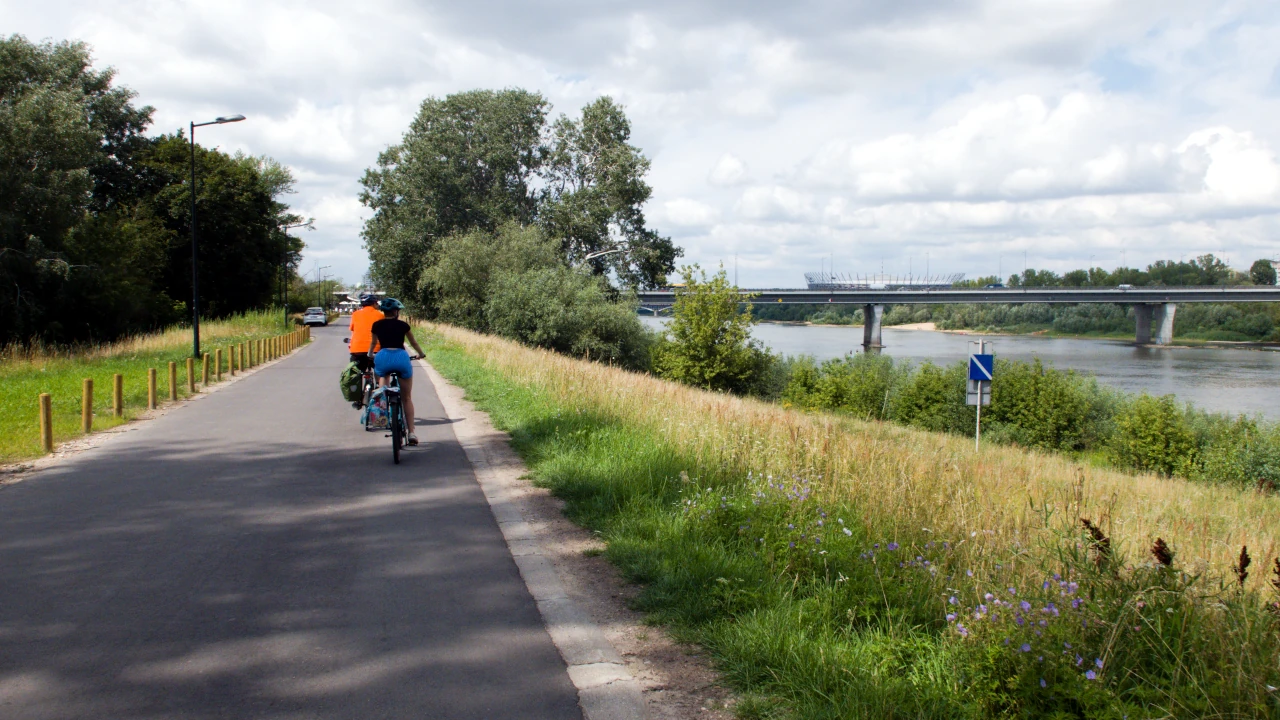 Dwoje rowerzystów poruszających się asfaltową drogą wzdłuż Wisły. W tle Most Łazienkowski.