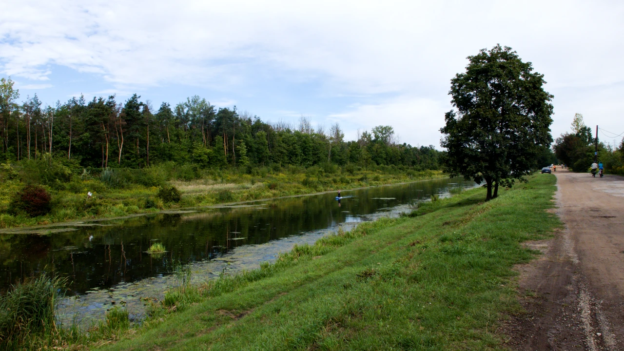 Po lewej stronie kanał biegnący między trawiastymi brzegami. Na wodzie ktoś płynie na supie. Po prawej stronie szutrowa droga z zaparkowanym samochodem i osobami na rowerach.