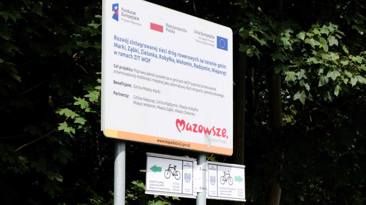 Tablica z informacją o sfinansowaniu inwestycji ze środków Unii Europejskiej. Poniżej drogowskazy wskazujące rowerowy dojazd do pobliskich miejscowości.