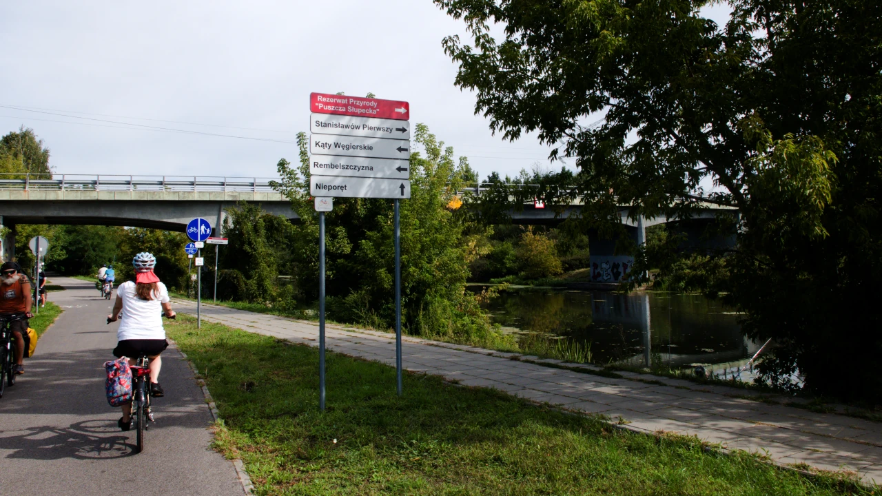 ilka osób na rowerach porusza się po drodze dla rowerów. Po prawej stronie tablice informacyjne z nazwami miejscowości i kierunkami jazdy. W tle most drogowy.