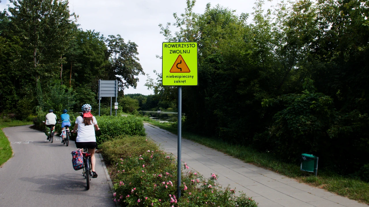 Kilka osób na rowerach poruszających się po drodze rowerowej. Po prawej tablica z napisem: rowerzysto zwolnij, niebezpieczny zakręt.
